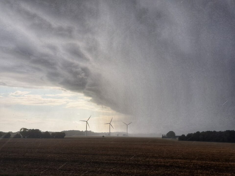 Storm clouds unleash a deluge of rain over the fields of Bornholm. Wind turbines turn on the horizon as they disappear into the grey. A hint of blue sky and white clouds still remain to the left of the shot.